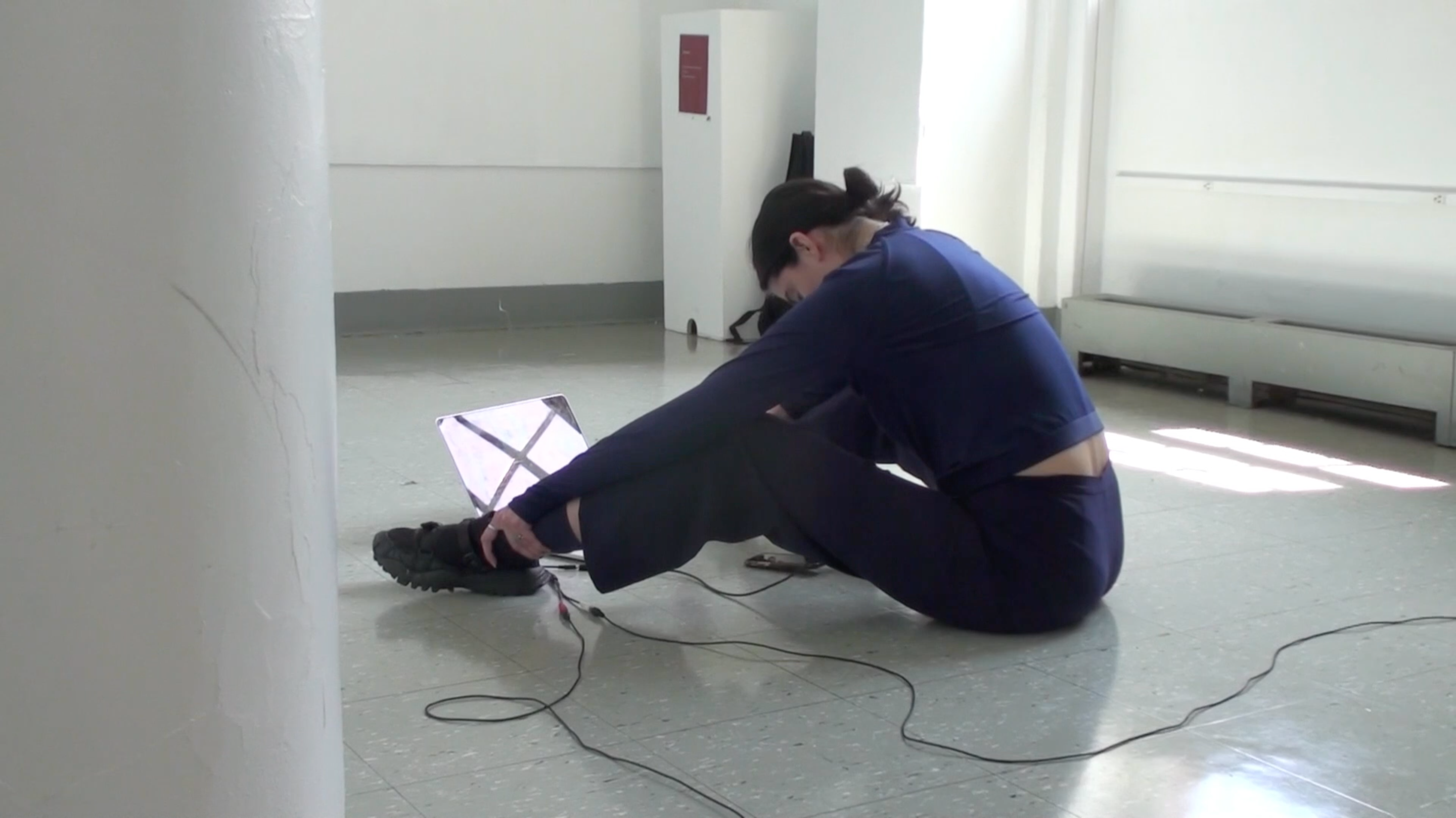 Performance still, body in an all blue fit sits with their legs open facing a computer, hands on ankles, head faces down towards the floor. Cables from the computer attach to their body.